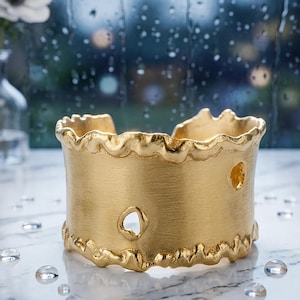 May include: A wide gold cuff bracelet with a brushed matte finish and an irregular, wavy top and bottom edge. The surface features two small, organic-shaped cutouts, one positioned near the center and one toward the right side.