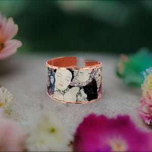 May include: A wide, open-band ring featuring a copper-toned metal finish. The exterior surface displays a detailed, multi-colored artistic print of a human face and abstract patterns in shades of gold, black, and soft pink, with a smooth edge.