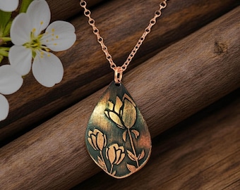 May include: A teardrop-shaped pendant necklace featuring an etched floral design of three tulips. The pendant has a dark, oxidized finish with copper-toned highlights on the petals and stems, suspended from a delicate copper-colored metal chain.