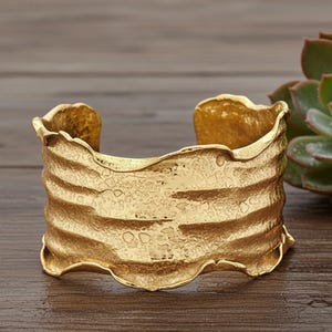 May include: A gold-colored cuff bracelet with a textured, organic design. The wide bracelet features a wavy, layered pattern and a rough, hammered surface. The open design allows for easy wear. This statement piece is a bold accessory.