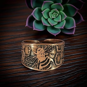May include: A wide copper-toned metal ring featuring an embossed design of a bee surrounded by a geometric honeycomb pattern. The band has an open back construction and a dark, antiqued finish that highlights the intricate details of the insect.