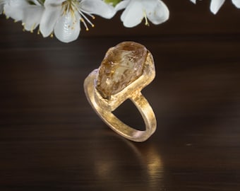 May include: A gold ring featuring a large, raw, amber-colored gemstone set in a textured, matte gold band. The ring is positioned upright, showcasing the irregular, faceted surface of the stone and the metallic finish of the band's setting.