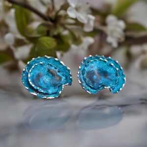 May include: A pair of stud earrings featuring a textured, circular design with a vibrant turquoise blue patina finish. The edges of the earrings have a metallic, scalloped appearance with subtle dark grey undertones visible within the centre.