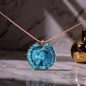 May include: A round pendant necklace featuring a textured, vibrant blue surface with metallic copper-toned edges. The pendant is suspended from a thin, matching copper-coloured snake chain, creating a distinct contrast between the blue and metallic tones.