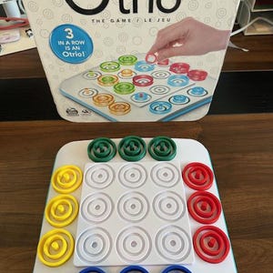 May include: A close-up of a game board with 16 white circles, each with a smaller circle in the center. There are four sets of colored rings, green, yellow, red, and blue, that can be placed on the board. The game is called "Otrio: The Game/Le Jeu".