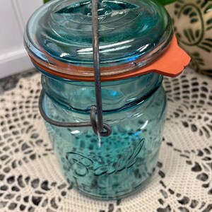 May include: A vintage blue glass Ball jar with a wire bail closure and a red rubber lid. The jar has a textured surface and the word "Ball" is embossed on the front.