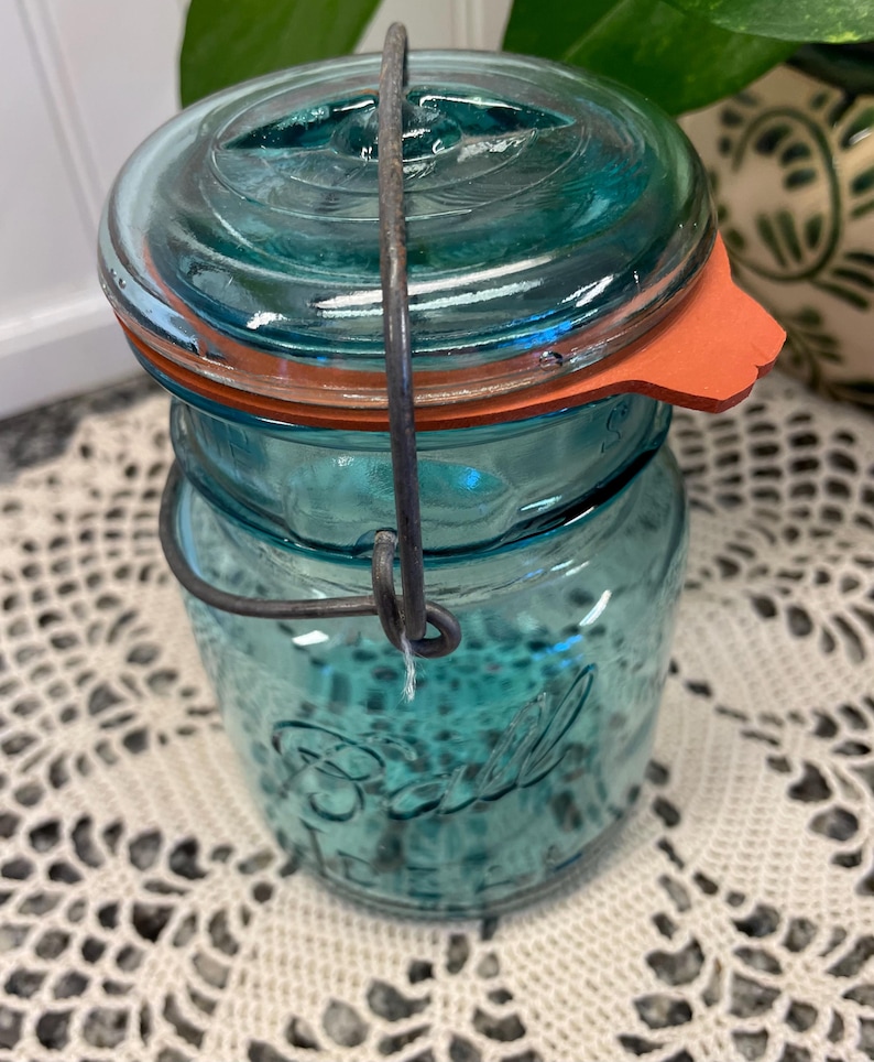 May include: A vintage blue glass Ball jar with a wire bail closure and a red rubber lid. The jar has a textured surface and the word "Ball" is embossed on the front.