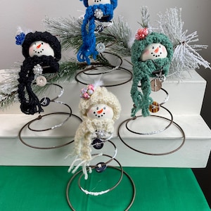 May include: Five handmade snowman ornaments with crocheted hats and scarves in various colors, including blue, black, green, and cream. Each snowman has a button nose and mouth, and is attached to a metal spring base. Decorative accents include buttons and faux pearls.