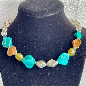 May include: A necklace with large, irregularly shaped beads in shades of turquoise, amber, and yellow-green. The necklace also features silver-toned and clear, round beads. The necklace is displayed on a black mannequin.