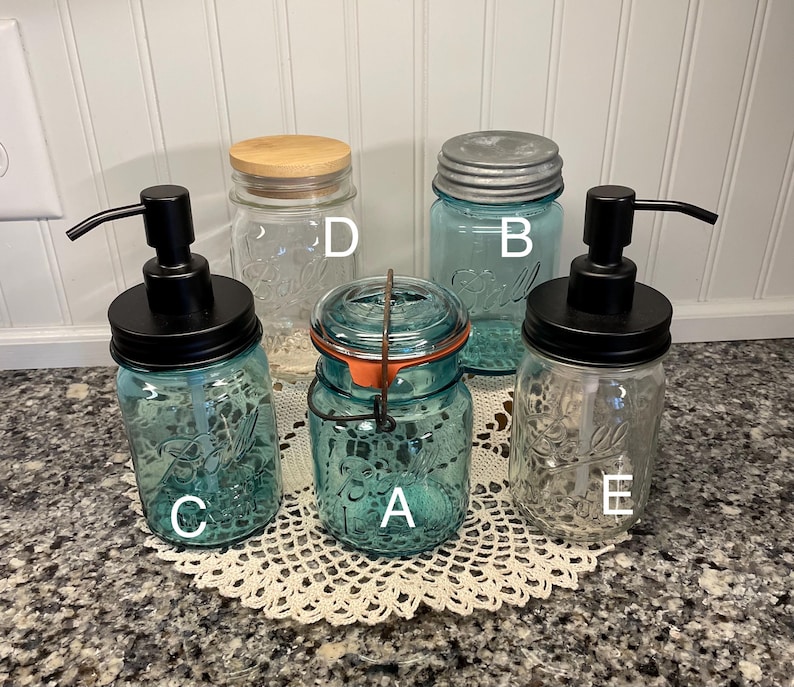 May include: Five glass jars with different lids and pumps. The jars are clear glass with a blue tint. The lids are a mix of wood, metal, and black plastic. The pumps are black plastic. The jars are labeled A, B, C, D, and E.