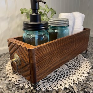 May include: A wooden box with decorative carvings and brass feet, used as a bathroom organizer. It contains a glass soap dispenser with a black pump, two glass jars with lids, and a white hand towel.