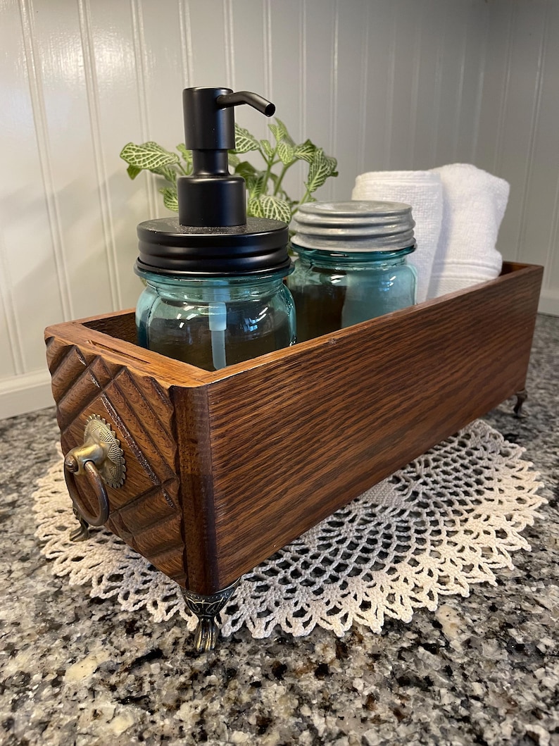 May include: A wooden box with decorative carvings and brass feet, used as a bathroom organizer. It contains a glass soap dispenser with a black pump, two glass jars with lids, and a white hand towel.