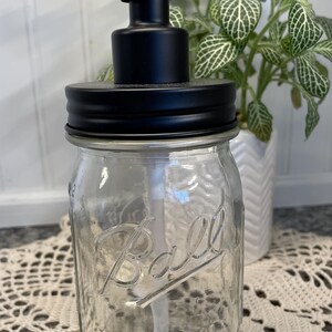 May include: Black soap dispenser pump attached to a clear glass Ball Mason jar with a black lid. The jar is sitting on a white crocheted doily.