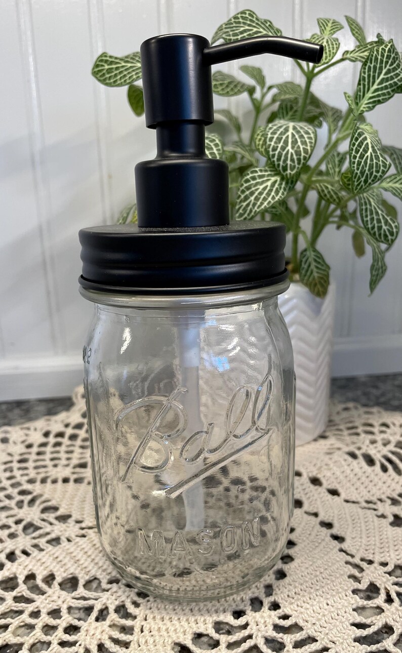 May include: Black soap dispenser pump attached to a clear glass Ball Mason jar with a black lid. The jar is sitting on a white crocheted doily.