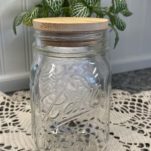 May include: A clear glass Ball Mason jar with a wooden lid. The jar is sitting on a white crocheted doily.