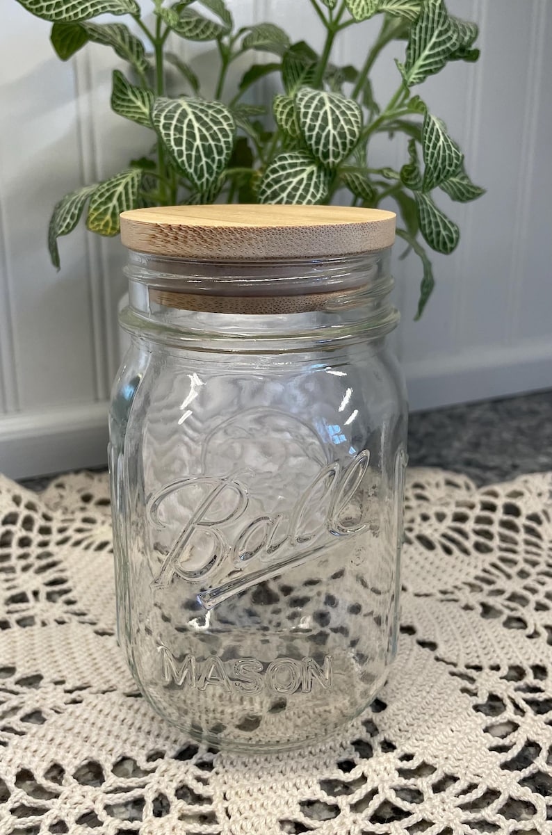 May include: A clear glass Ball Mason jar with a wooden lid. The jar is sitting on a white crocheted doily.