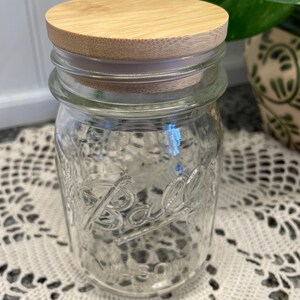 May include: A clear glass Ball mason jar with a wooden lid. The jar is sitting on a white crocheted doily.