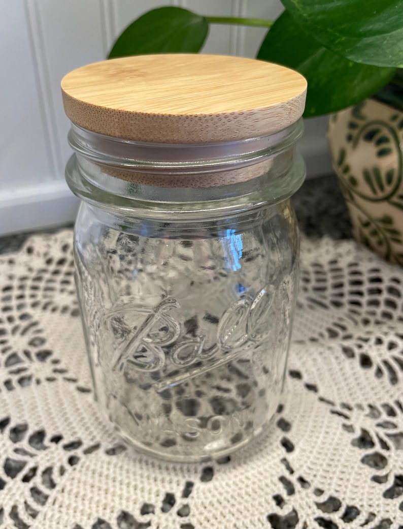 May include: A clear glass Ball mason jar with a wooden lid. The jar is sitting on a white crocheted doily.