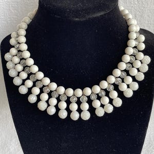May include: A white beaded necklace with multiple strands. The necklace features round and floral-shaped beads, interspersed with clear, faceted beads. The necklace is displayed on a black jewelry bust.