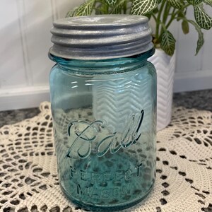 May include: A vintage blue Ball Perfect Mason jar with a silver lid. The jar is sitting on a white crocheted doily.