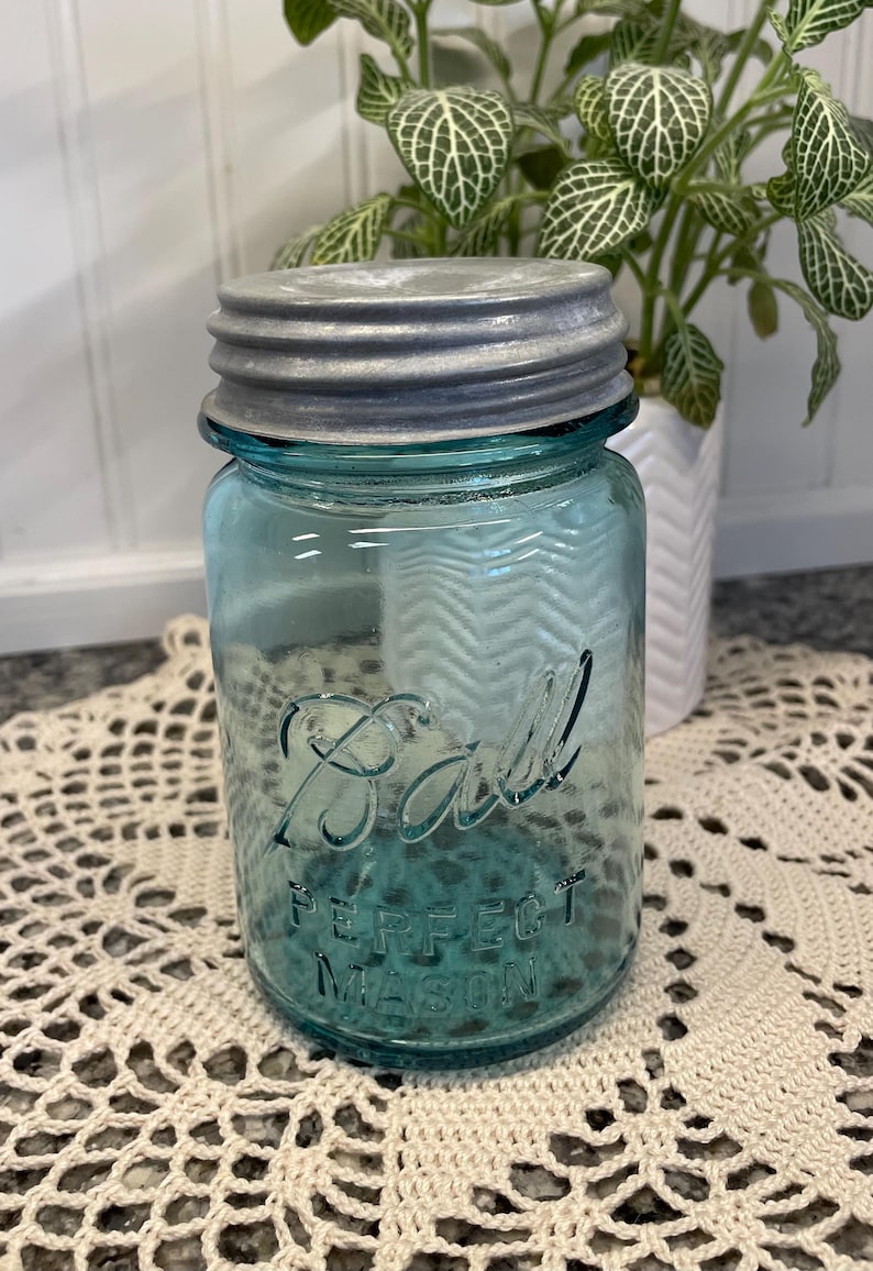 May include: A vintage blue Ball Perfect Mason jar with a silver lid. The jar is sitting on a white crocheted doily.