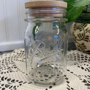 May include: A clear glass Ball Mason jar with a wooden lid. The jar is empty and sitting on a white crocheted doily.