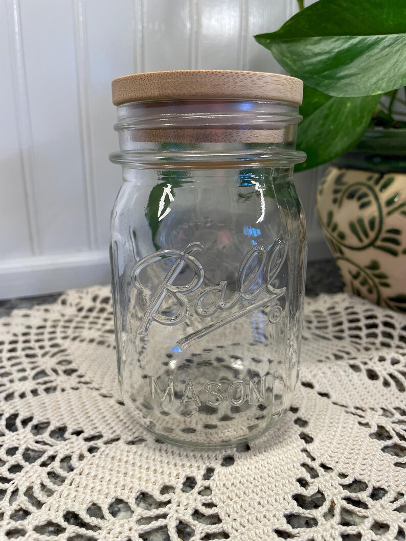May include: A clear glass Ball Mason jar with a wooden lid. The jar is empty and sitting on a white crocheted doily.