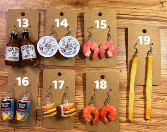 May include: A collection of novelty earrings, including miniature bottles of barbecue sauce, Cool Whip containers, shrimp, french fries, Minute Maid cans, and cheeseburgers. Each pair is attached to a brown card with a number.