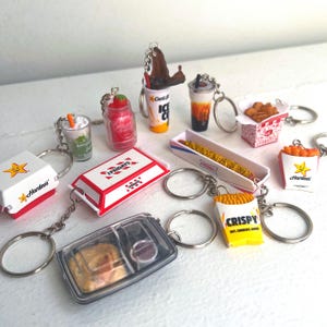 Puede incluir: Una colección de llaveros de comida en miniatura, que incluyen una caja de Hardee's, una caja de Friday's, un recipiente transparente con una hamburguesa y varias tazas de bebidas. Otros llaveros presentan papas fritas, un perrito caliente y un tarro de fresas.