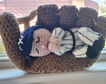 May include: A crocheted baseball mitt with a small crocheted baseball player sleeping inside. The mitt is brown and the player is wearing a white and black striped uniform with a blue hat.
