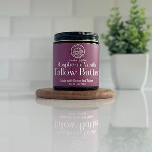 May include: A jar of Raspberry Vanilla Tallow Butter with a black lid and a purple label. The label reads "Limited Edition" and "Made with Grass-fed Tallow." The jar is on a small wooden coaster. Net weight is 4 oz (113 g).