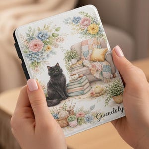 May include: A white tablet case featuring a watercolor illustration of a black cat, books, a cozy armchair, and floral accents. The case is held by a person. The name "Gwendoly" is written on the case.