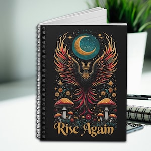 May include: A black spiral notebook with a colorful illustration of a phoenix rising from a bed of mushrooms and flowers. The moon is in the background and the text "Rise Again" is at the bottom.