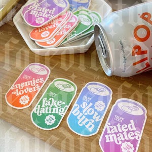 May include: Four colorful can shaped stickers with different slogans. The stickers are on a wooden surface. The slogans are "enemies to lovers", "fake dating", "slow burn", and "rated mates".