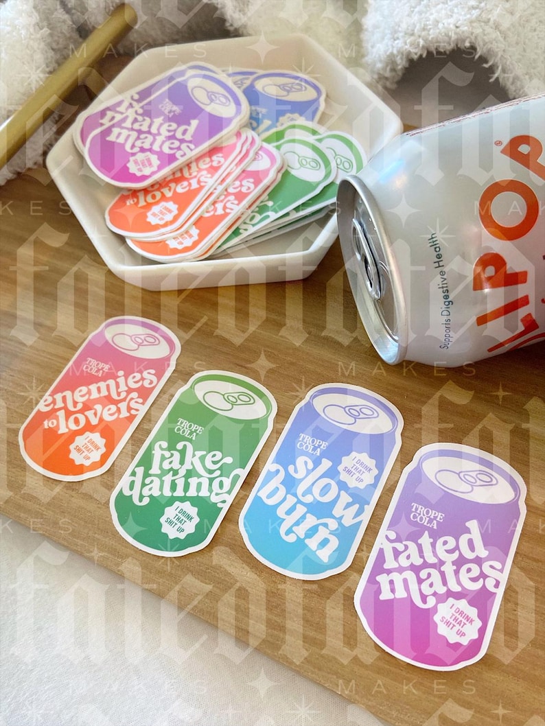 May include: Four colorful can shaped stickers with different slogans. The stickers are on a wooden surface. The slogans are "enemies to lovers", "fake dating", "slow burn", and "rated mates".