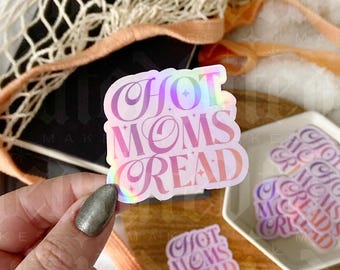 Puede incluir: Una pegatina holográfica con el texto "Hot Moms Read" en letras rosa y blancas. La pegatina está sobre una superficie de madera con un plato de cerámica blanco y una bolsa de malla.