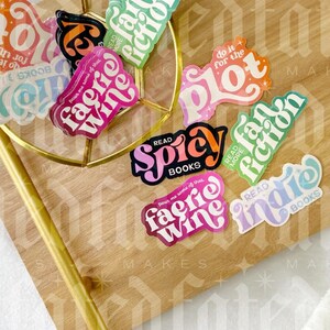 May include: A collection of colorful stickers with book-themed phrases like "pour me some of that faerie wine", "do it for the plot", "read more indie", and "read more spicy".