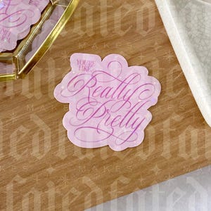 Op de afbeelding: Een roze sticker met de woorden "You're Like Really Pretty" in een cursief lettertype. De sticker heeft een lichtroze rand en ligt op een houten oppervlak. Extra stickers zijn zichtbaar op de achtergrond.