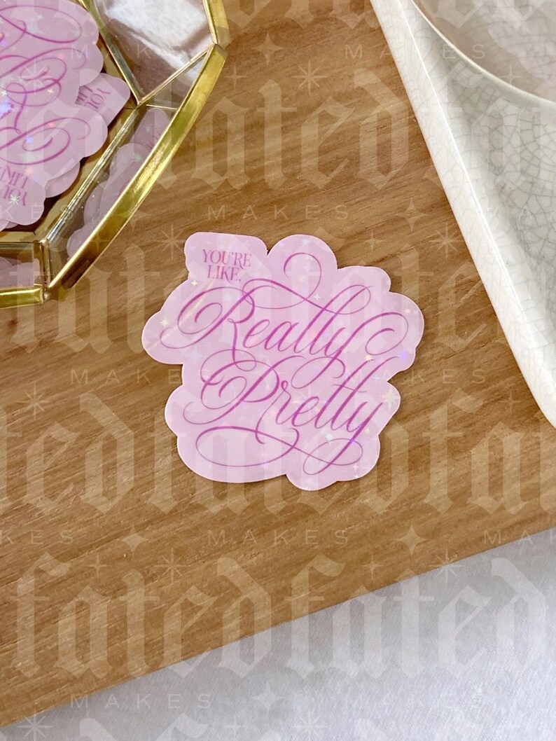Op de afbeelding: Een roze sticker met de woorden "You're Like Really Pretty" in een cursief lettertype. De sticker heeft een lichtroze rand en ligt op een houten oppervlak. Extra stickers zijn zichtbaar op de achtergrond.