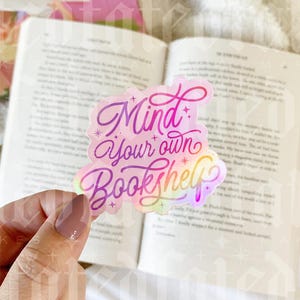 Peut inclure: Un autocollant holographique avec un dégradé rose, violet et bleu. L'autocollant dit "Mind your own Bookshelf" dans une police de caractères fantaisiste.
