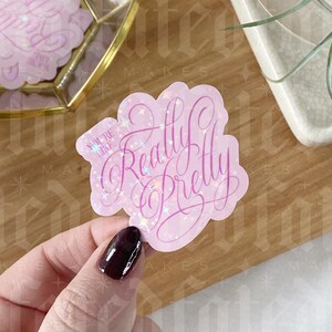 Op de afbeelding: Een roze sticker met de tekst "You're Like Really Pretty" in een cursief lettertype. De sticker heeft een holografisch glittereffect. Een hand met donker bordeauxrode nagellak houdt de sticker vast.