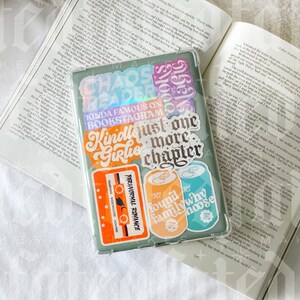 Pu&ograve; includere: Una custodia per telefono verde con vari adesivi colorati. Gli adesivi presentano frasi come "Chaos Readers", "Kinda Famous on Bookstagram", "Just One More Chapter" e "Found Family Why Moose".