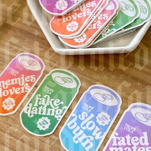 May include: Four colorful stickers shaped like soda cans with different phrases on them. The phrases are "Fated Mates", "Fake Dating", "Slow Burn", and "Enemies Lovers". Each sticker has a white circle with the text "I Drink That Shit Up" in black.
