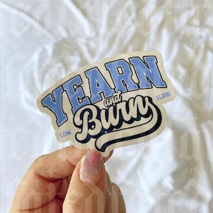 May include: A sticker with the text "YEARN and Burn" in a blue and black design. The word "YEARN" is in a light blue, blocky font, and "Burn" is in a cursive font. The sticker has a white background and is held by a hand.