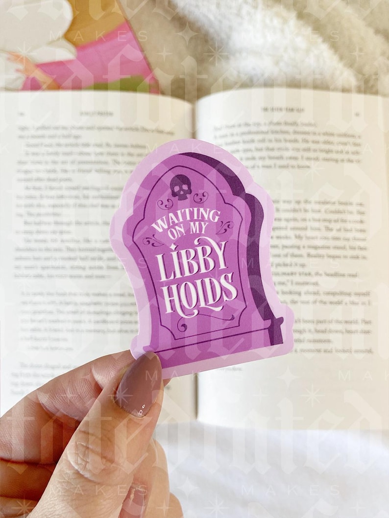 May include: Purple sticker shaped like a tombstone with a skull and crossbones at the top. The sticker says "Waiting on my Libby Holds".
