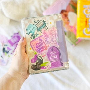 May include: A clear plastic case with a purple and white checkered pattern. The case has several stickers on it, including a book with the words "By Day" and "By Night", a purple eggplant, a black cat, and a tombstone with the words "Waiting on my Libby Hoids".