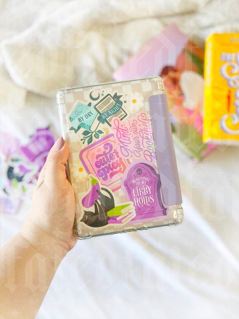 May include: A clear plastic case with a purple and white checkered pattern. The case has several stickers on it, including a book with the words "By Day" and "By Night", a purple eggplant, a black cat, and a tombstone with the words "Waiting on my Libby Hoids".