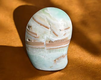 May include: A polished, rounded stone with a blend of pale green, white, and tan hues. The stone features layered bands and natural patterns, creating a unique aesthetic. The smooth surface reflects light, highlighting the stone's natural beauty.