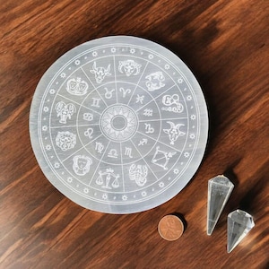 May include: A white selenite charging plate featuring a zodiac wheel design. The plate is surrounded by a circle of small stars. Two clear quartz crystal points are also shown.