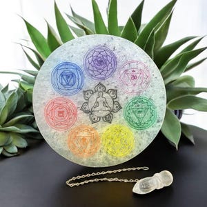 May include: A white circular crystal with seven colorful chakra symbols and a meditating figure in the center. A clear quartz pendulum with a silver chain is also pictured.